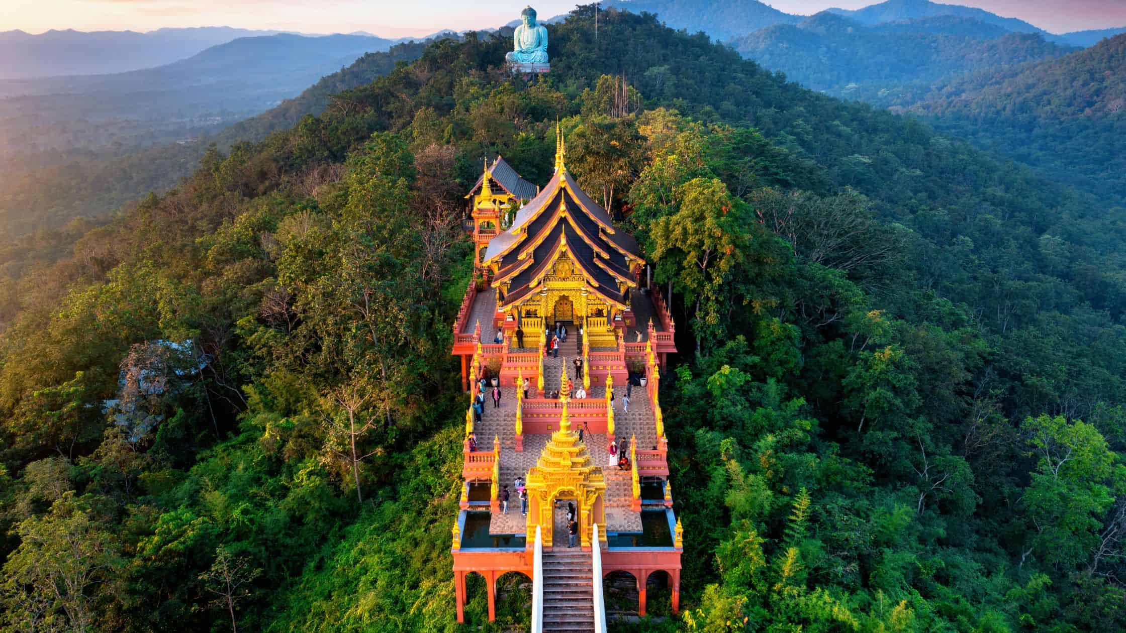 Aerial view of an ornate red-and-gold hilltop temple in the jungle mountains of northern Thailand with a large Buddha statue on a distant peak
